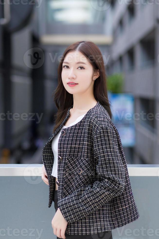 Portrait of a beautiful, long-haired Asian woman in a black pattern coat with braces on teeth standing and smiling outdoors in the city. photo