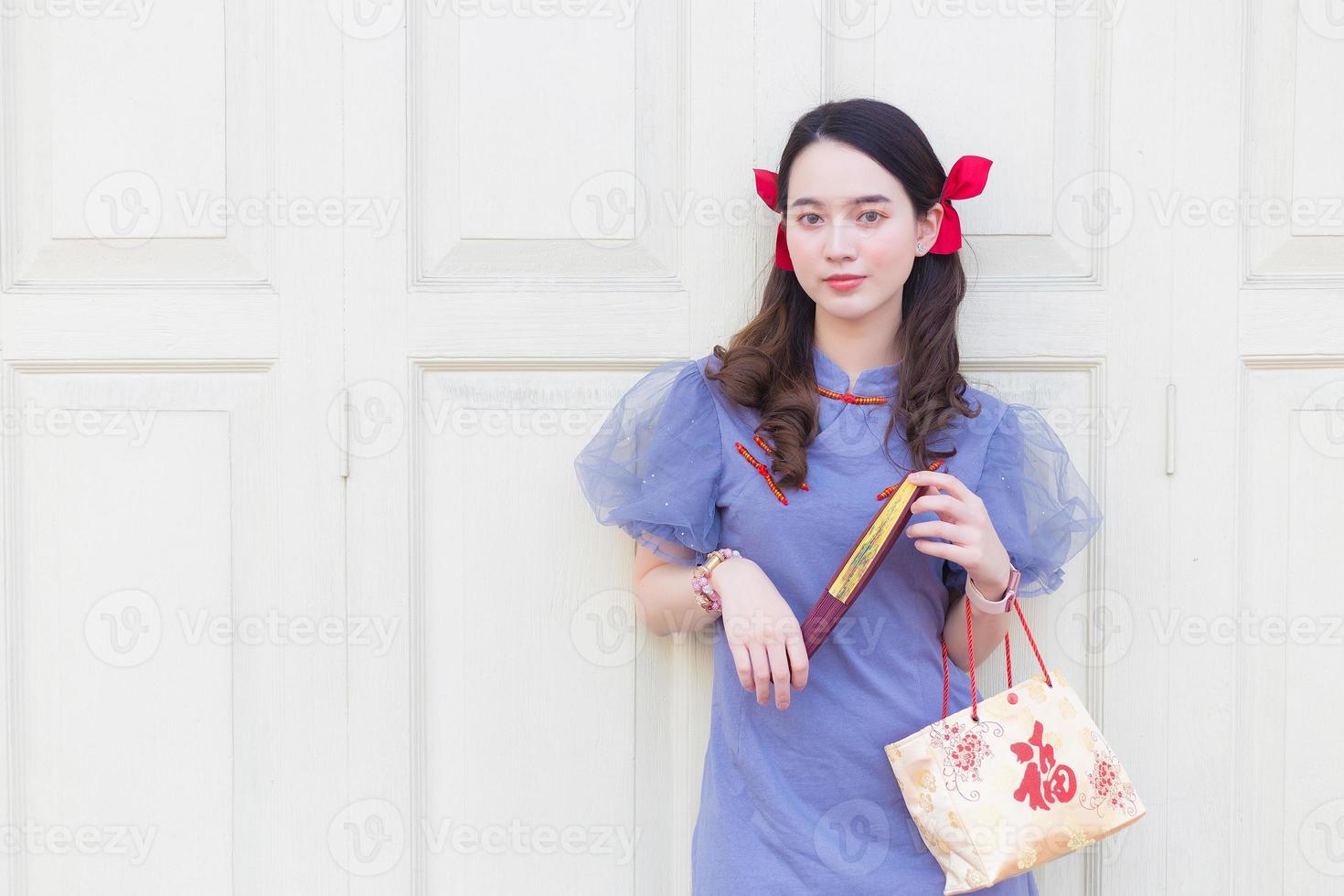 una mujer asiática vestida de qipao chino gris azulado sostiene la bolsa de tela que proyecta la palabra que significa feliz en chino y se para en una puerta de madera blanca como fondo. foto