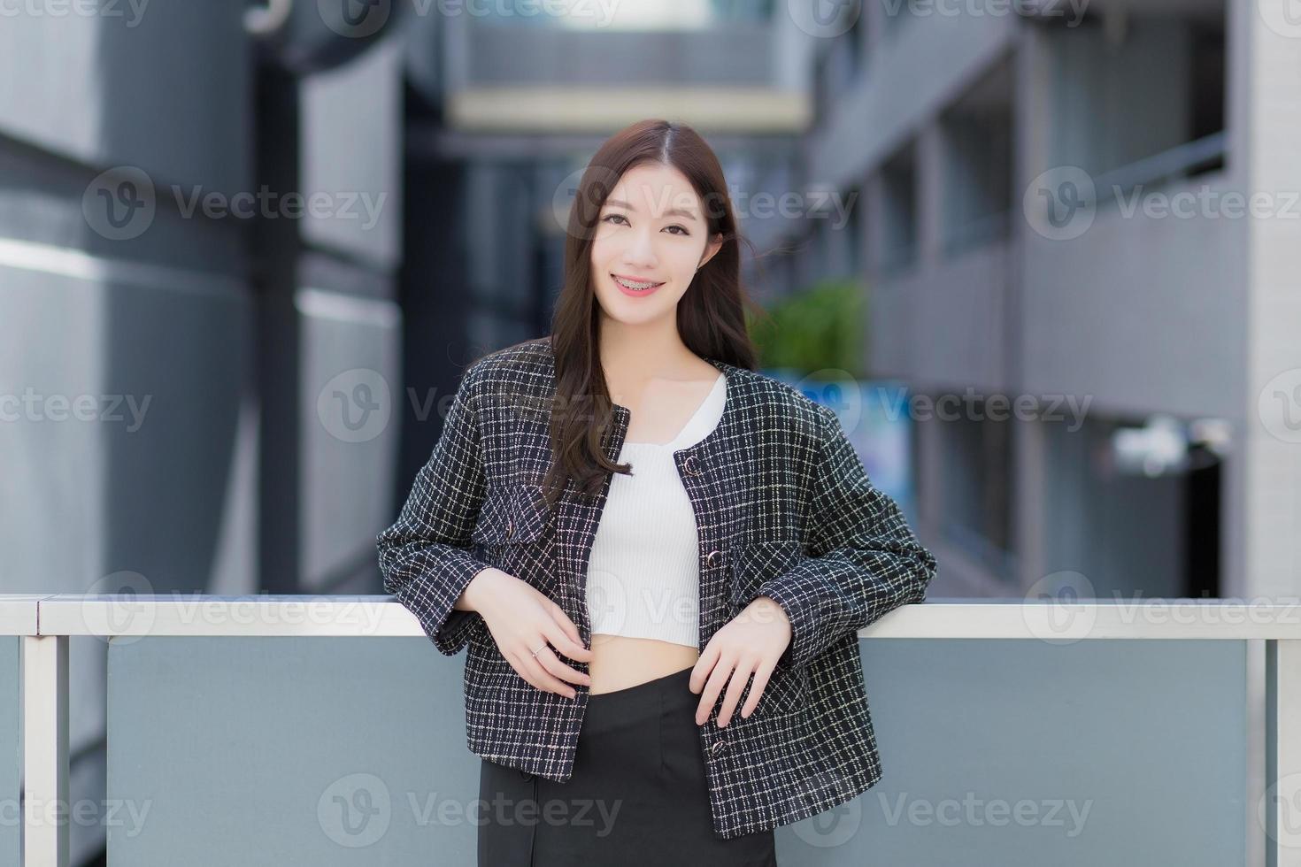 Portrait of a beautiful, long-haired Asian female in a black pattern coat with braces on teeth standing and smiling outdoors in the city. photo