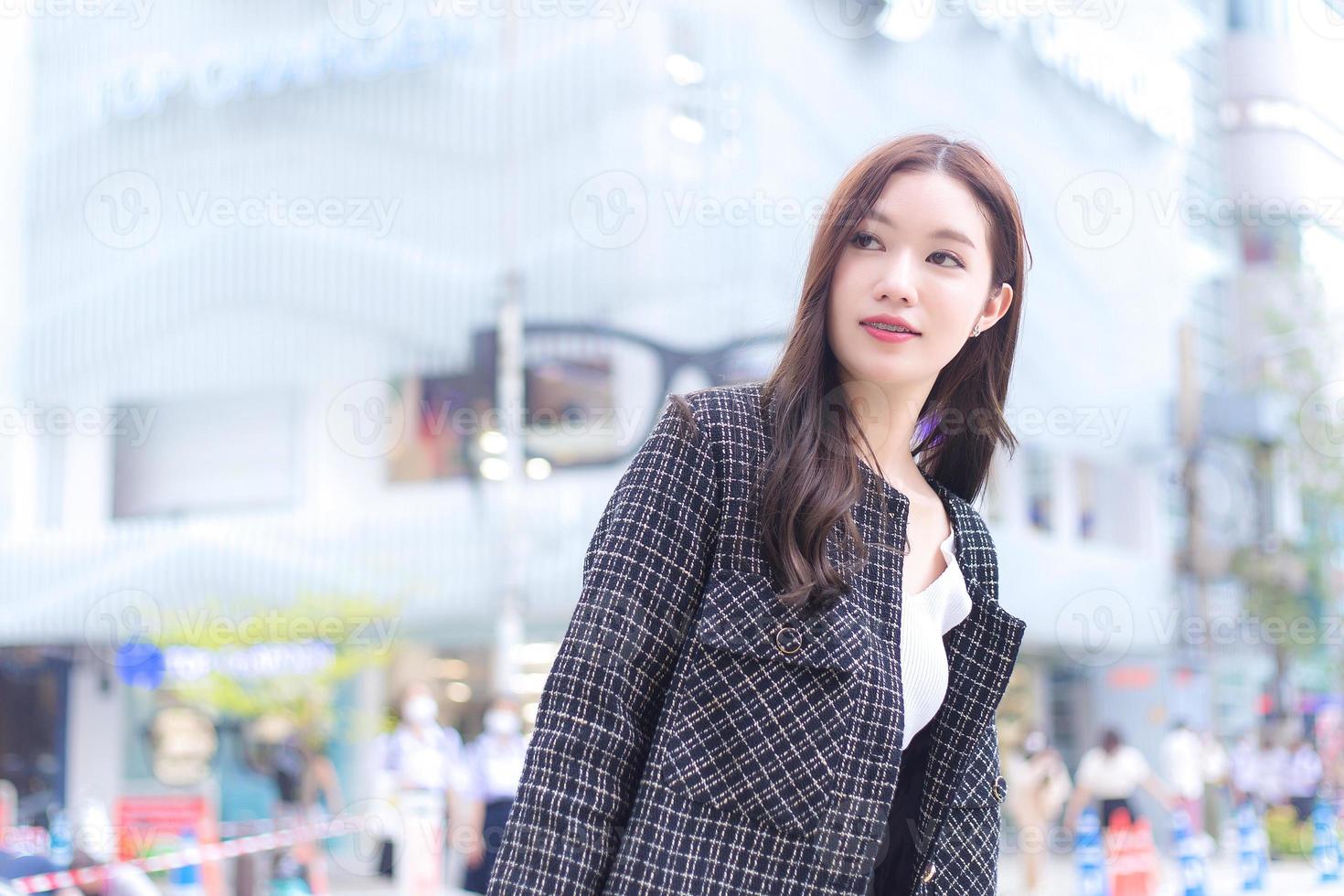 Portrait of a beautiful, long-haired Asian female in a black pattern coat with braces on teeth walking and smiling outdoors with the city the background.. photo
