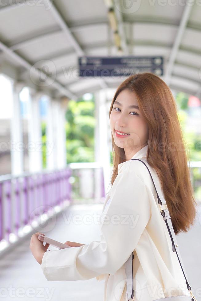 Beautiful young Asian working woman in white suit is out to meet a client at the office in a happy mood with a city background. photo