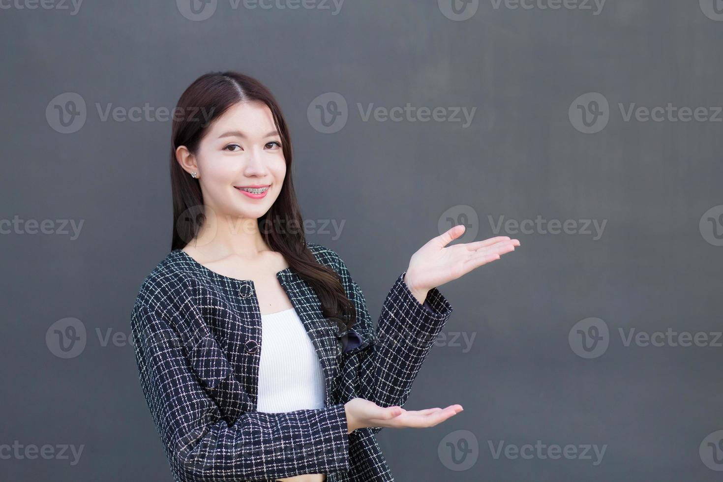 Asian professional working woman who wears black suit with braces on teeth is pointing hand to present something on the dark gray wall. photo