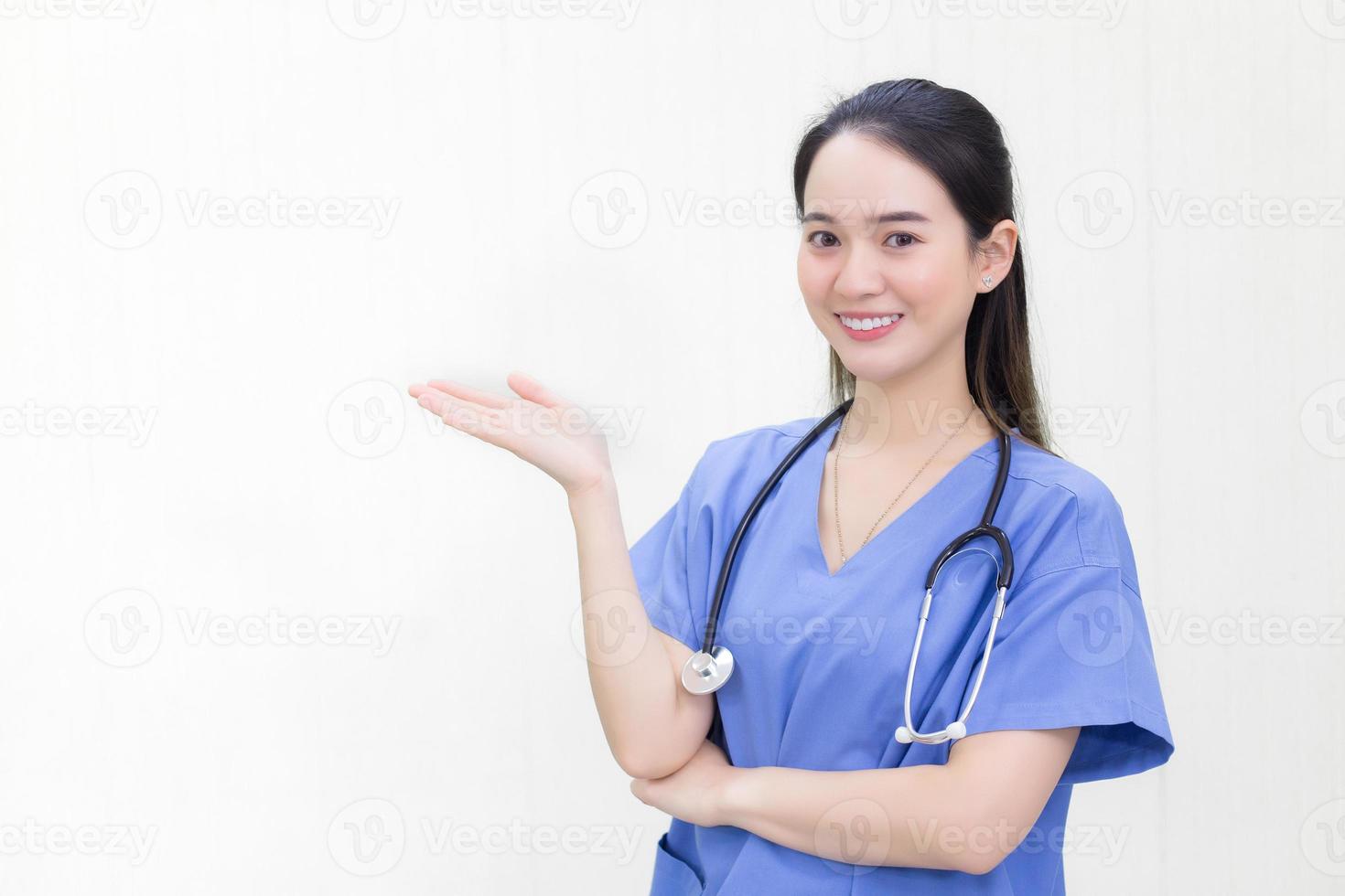 A beautiful young Asian woman doctor in a blue uniform stands and smiles while pointing to the top on a white background. photo