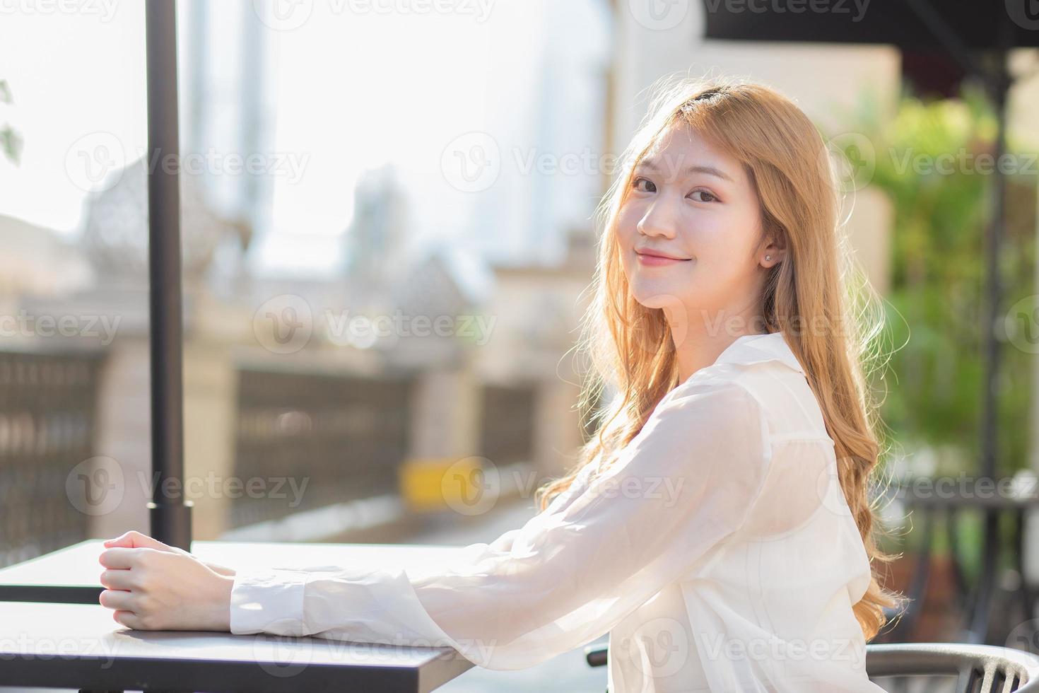 una hermosa mujer asiática con una camisa blanca de manga larga está sentada en una silla mirando hacia afuera. frente a una cafetería en una mañana soleada. foto