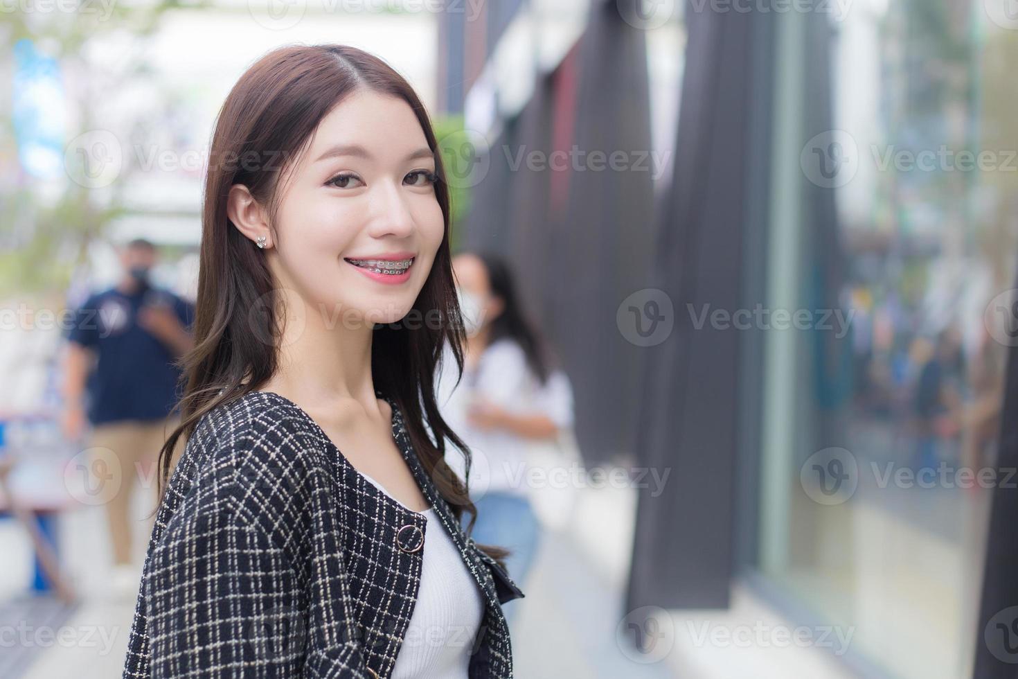 Portrait of a beautiful, long-haired Asian woman in a black pattern coat with braces on teeth walking and smiling outdoors in the city. photo