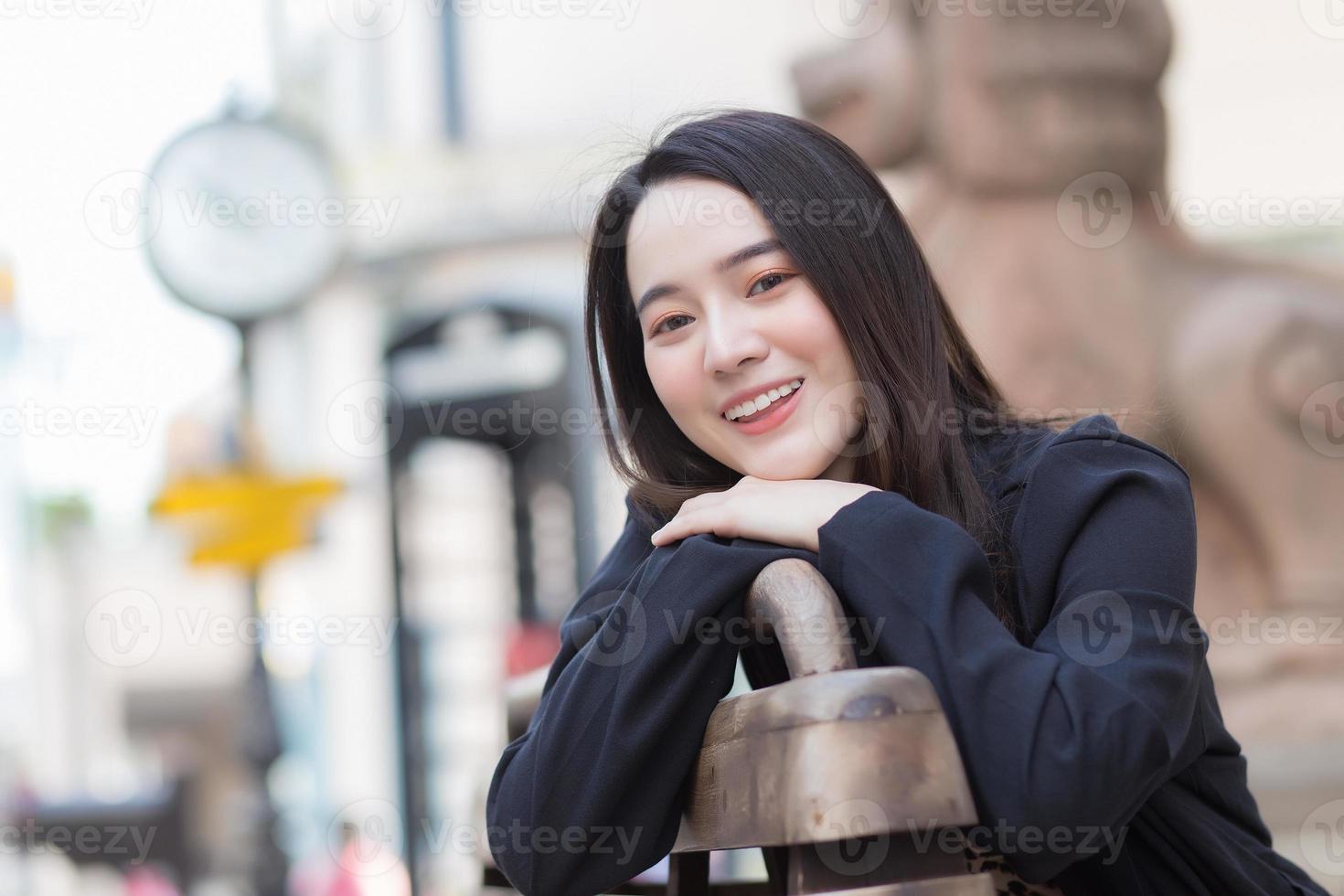 A beautiful Asian woman in a black long sleeve shirt sits happily on a chair in an outdoor park. photo