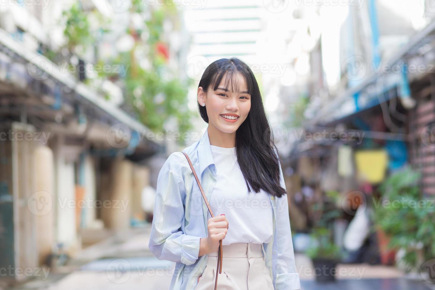 una joven asiática segura de sí misma que usa una camisa blanca azul y una bolsa sonríe alegremente y mira la cámara mientras viaja al trabajo por el casco antiguo. foto