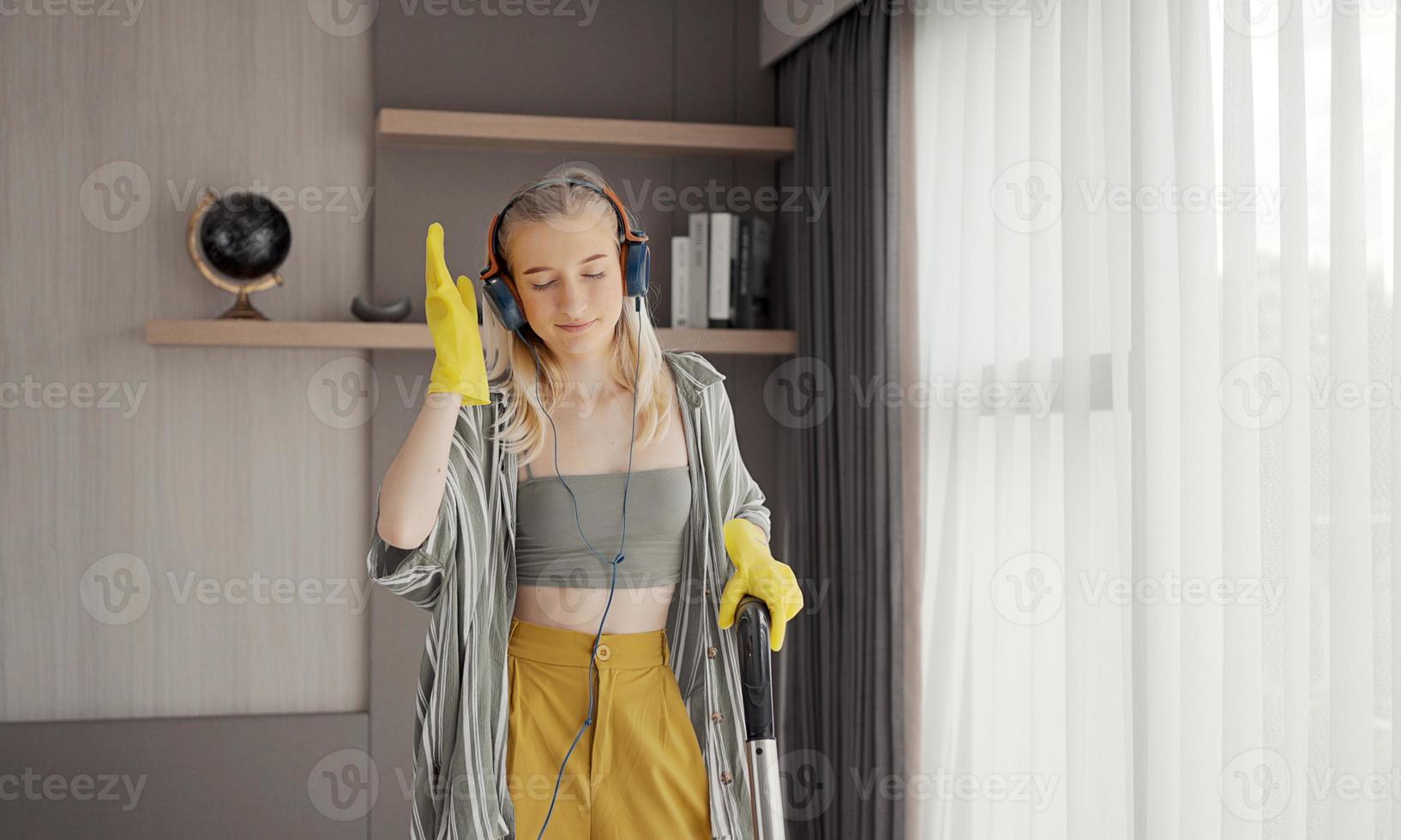 Young girl havig fun while cleaning floor with vacuum cleaner. Happy woman doing housework at home enjoy music wearing earphones. photo