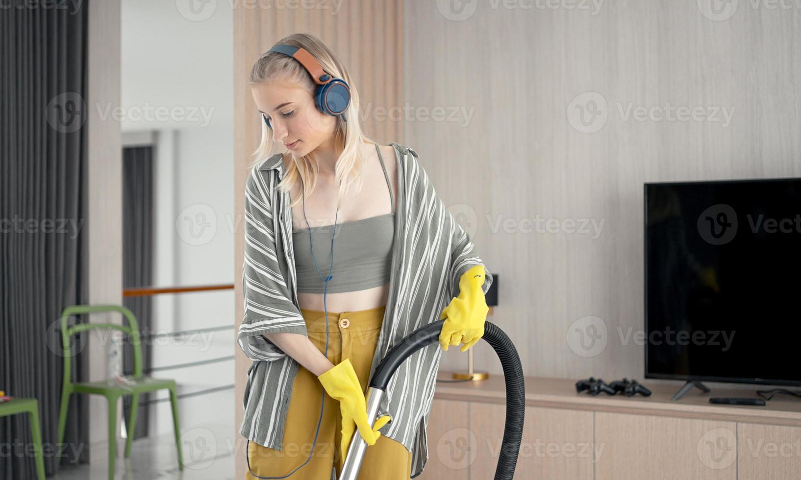 Young girl havig fun while cleaning floor with vacuum cleaner. Happy woman doing housework at home enjoy music wearing earphones. photo