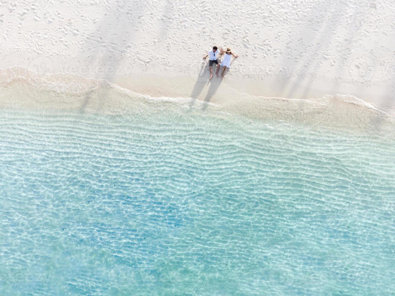 Young couple traveler sitting and relaxing at beautiful tropical white sand beach with wave foam and transparent sea, Summer vacation and Travel background Top view from drone photo