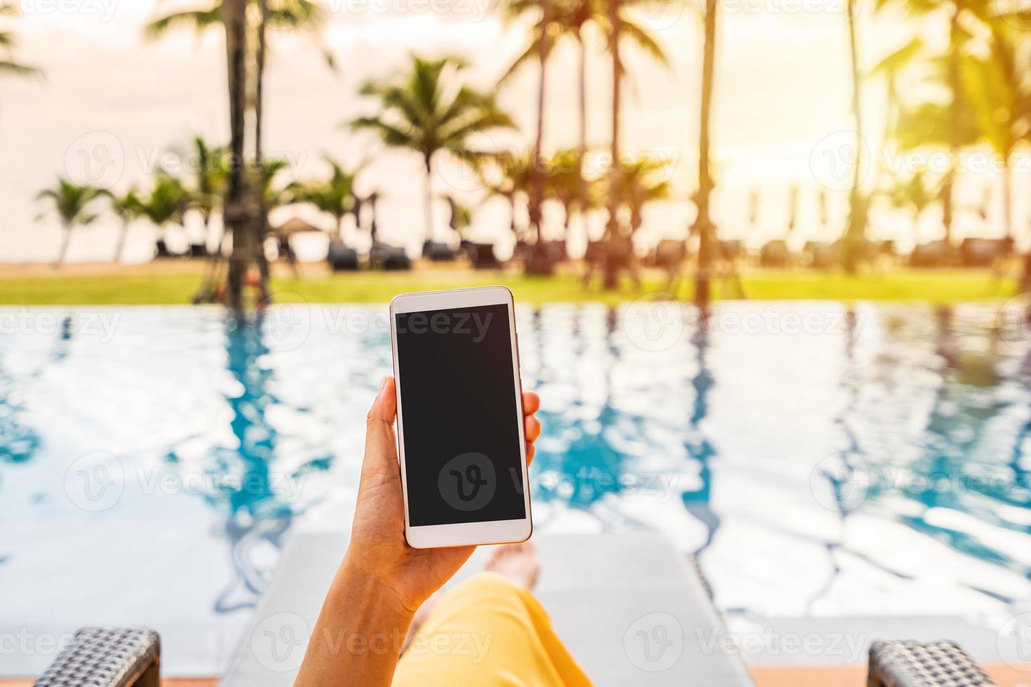 Young woman traveler relaxing and using a mobile phone by a hotel pool while traveling for summer vacation photo