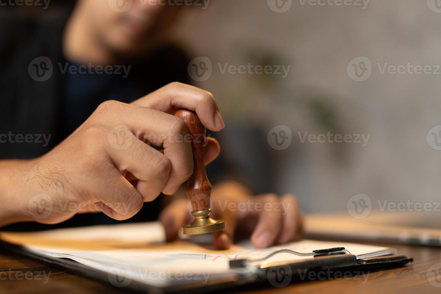 Close-up of a person's hand stamping with approved stamp on certificate document public paper at desk, notary or business people work from home, isolated for coronavirus COVID-19 protection photo