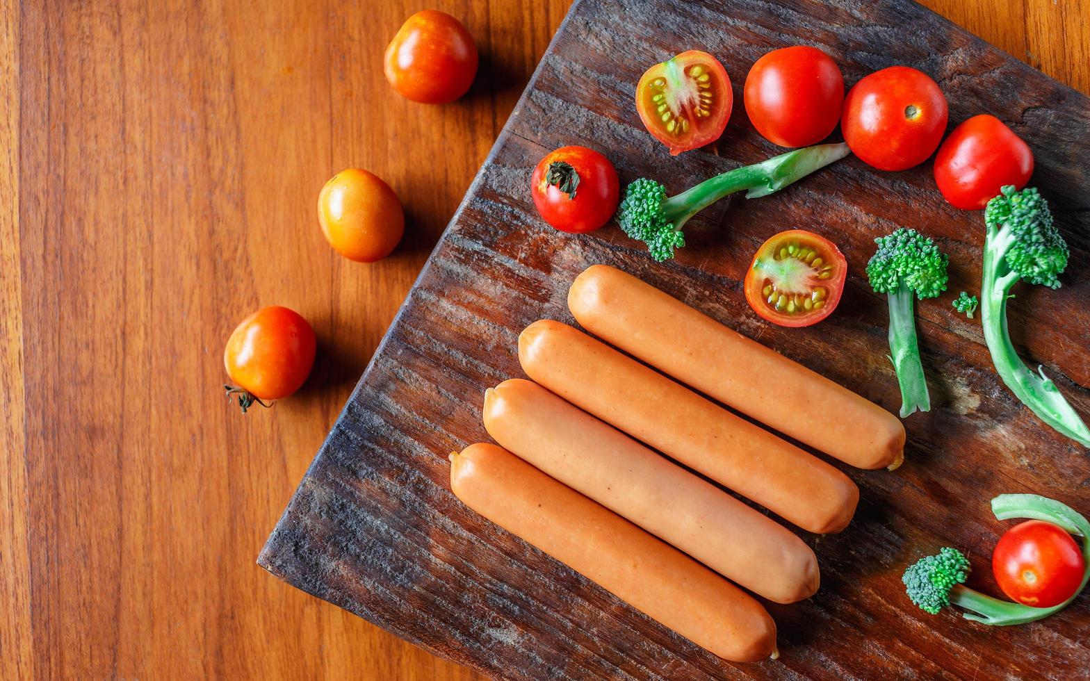 salchicha cruda en una tabla de cortar de madera con brócoli y tomates foto