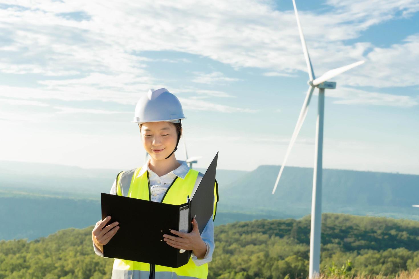 Beautiful caucasian woman in white helmet looking through some documents on clipboard while standing on farm with windmills. photo