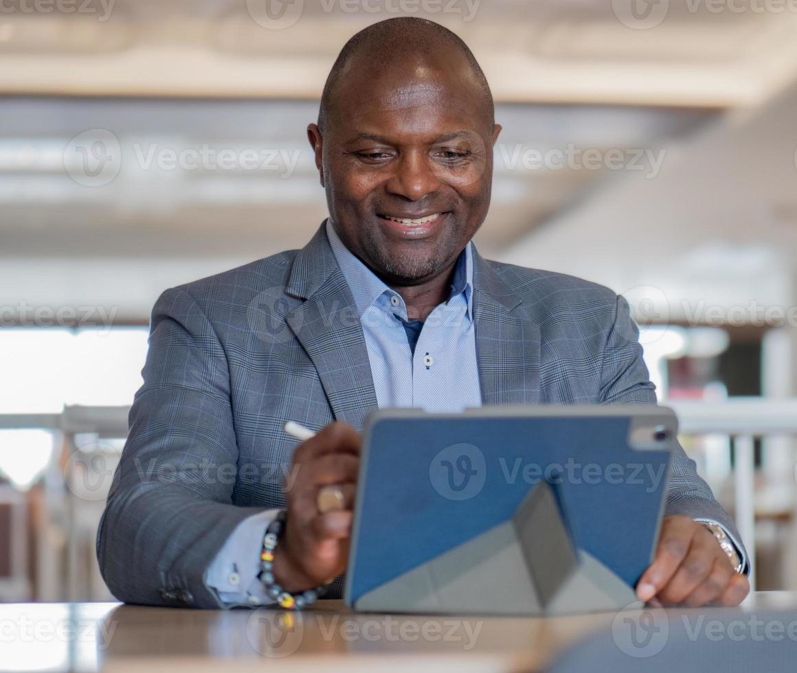 Portrait of happy black businessman in suit sitting at desk in modern office using tablet computer for remote meeting. Smiling black manager, successful working as white collar worker in corporate. photo