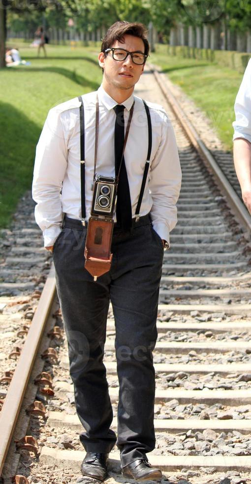 Young man wearing glasses and suspenders walking along a railway track with a vintage camera around his neck photo