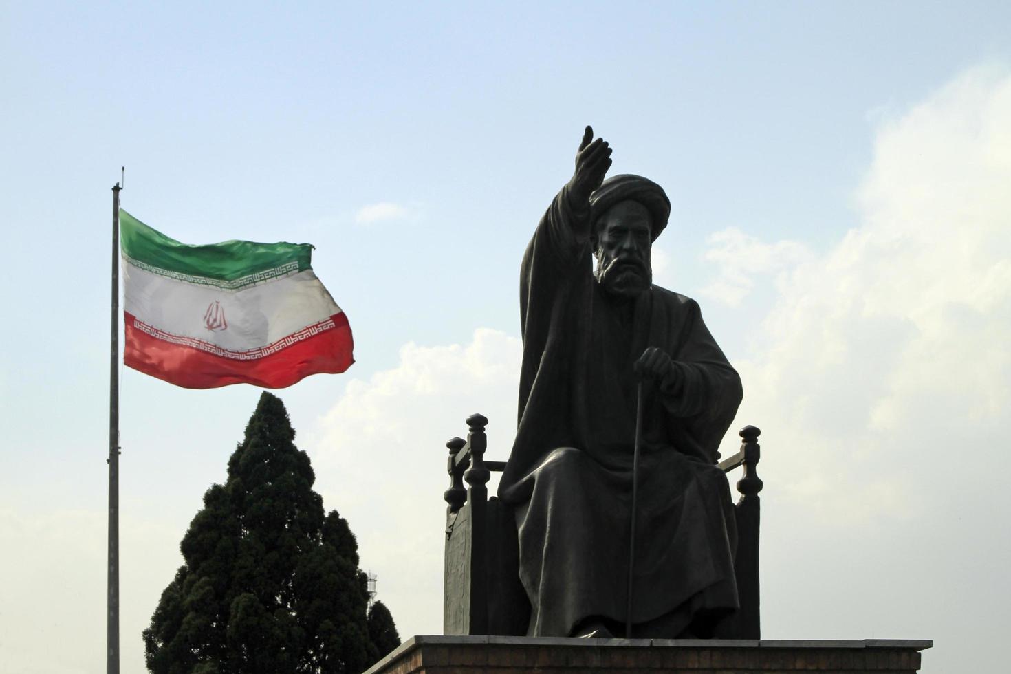 Tehran, Iran - June 10, 2018 - A large Iranian flag waving in the wind behind a statue of Khomeini in Tehran, Iran photo