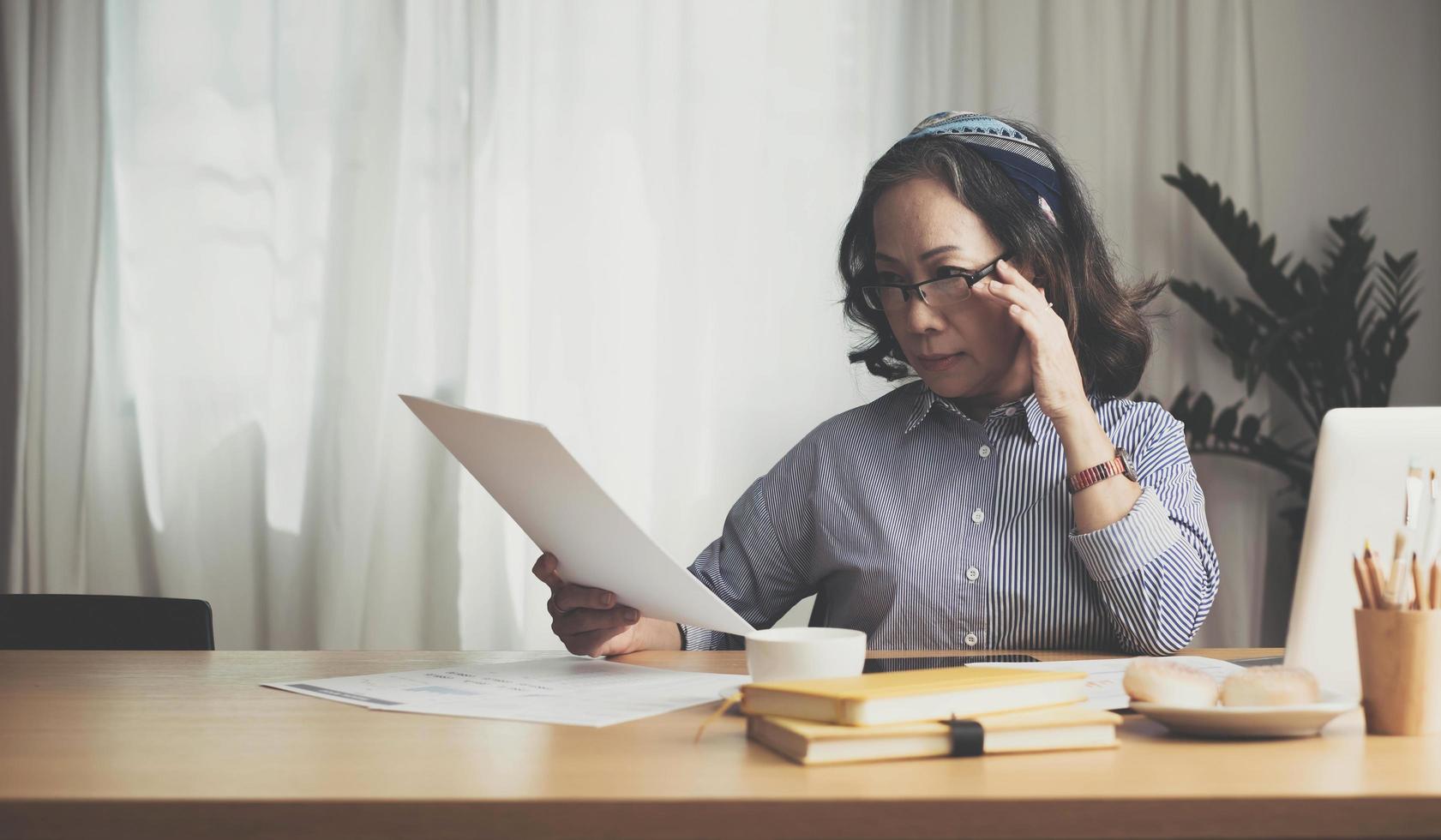 Seriously senior woman tworking at home, looking at laptop and waving paper correspondence, sitting at home, free space. photo