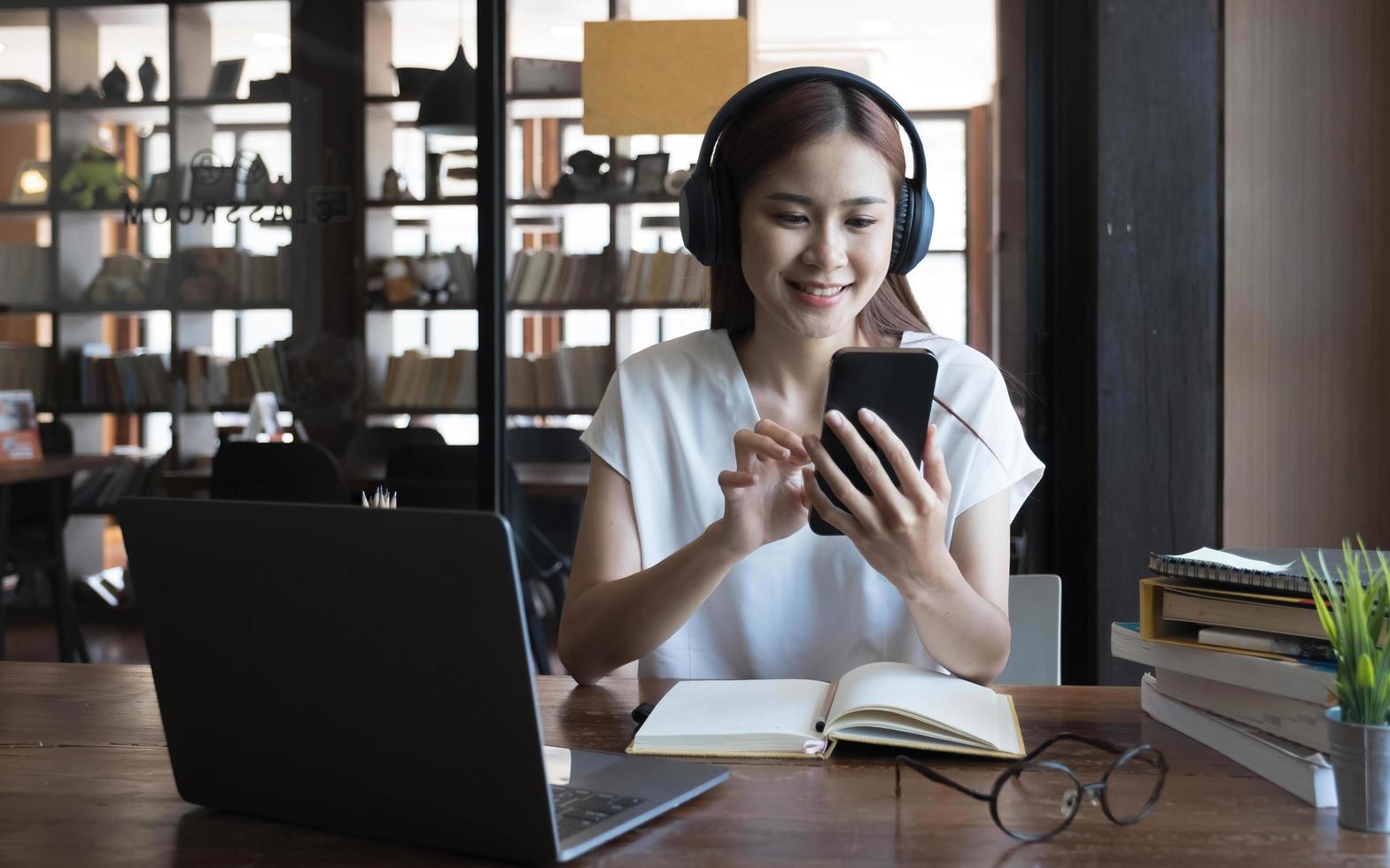 Beautiful millennial Asian female university student listening to music with her modern wireless headphones and using smartphone. photo