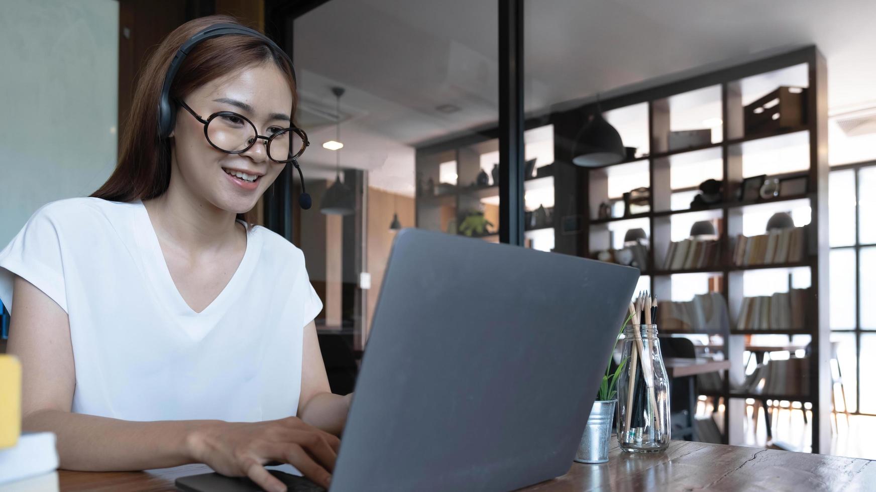 Smiling Asian young female using headset looking at laptop screen listen and learning online courses. Happy chinese business woman with headphones video call for customer service photo