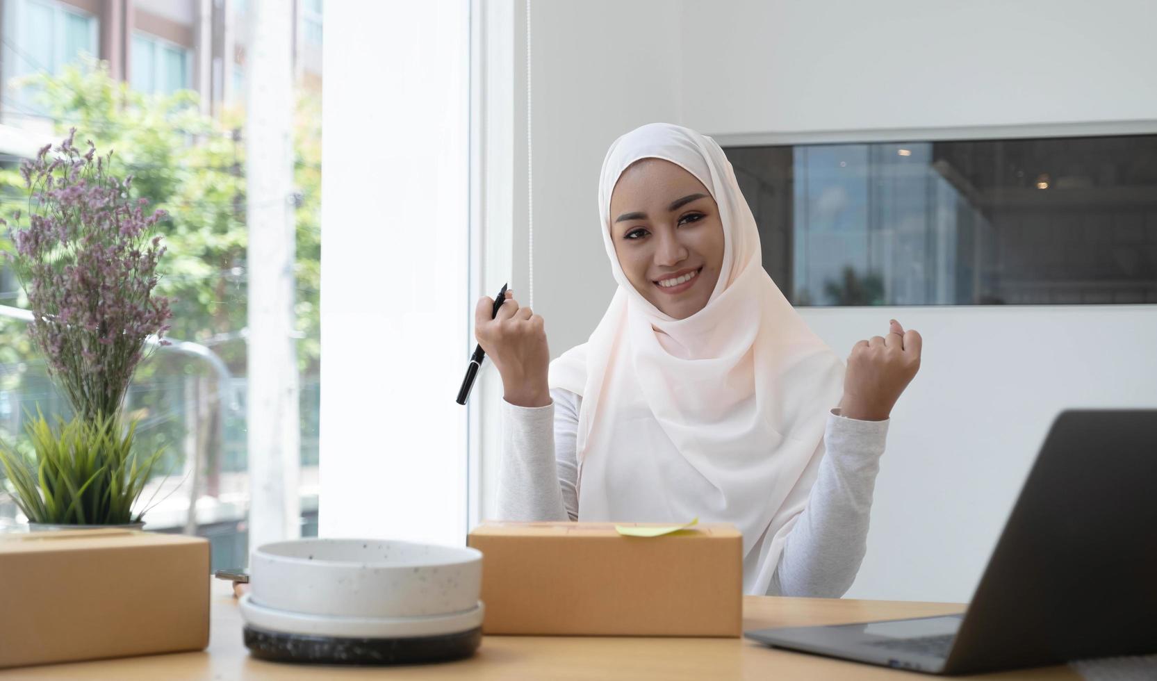 Portrait of a cute smiling Muslim woman wearing a light pink hijab sitting on an office chair. Looking at the camera is delighted while being able to sell online and the background of the box. photo
