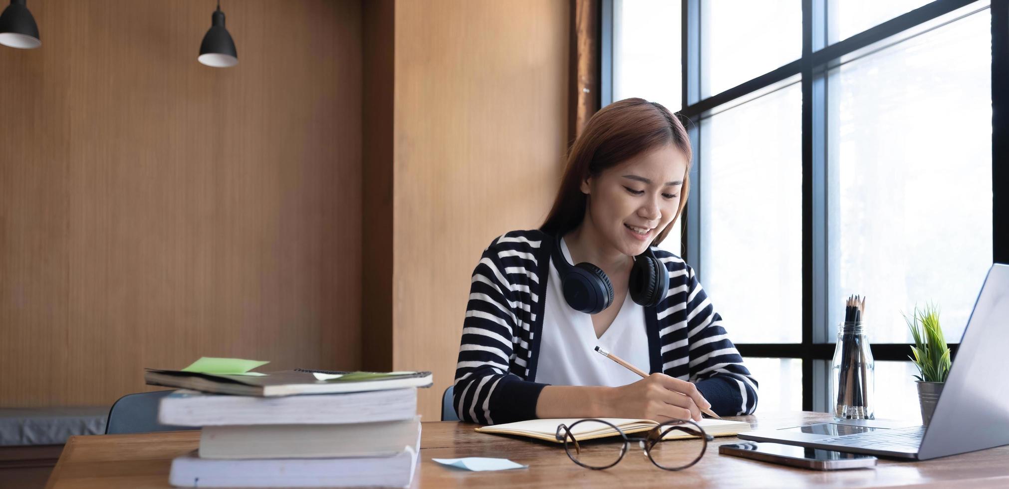 Smiling asian undergraduate teen girl student study in library with laptop books doing online research for coursework, making notes for essay homework assignment, online education e-learning concept photo