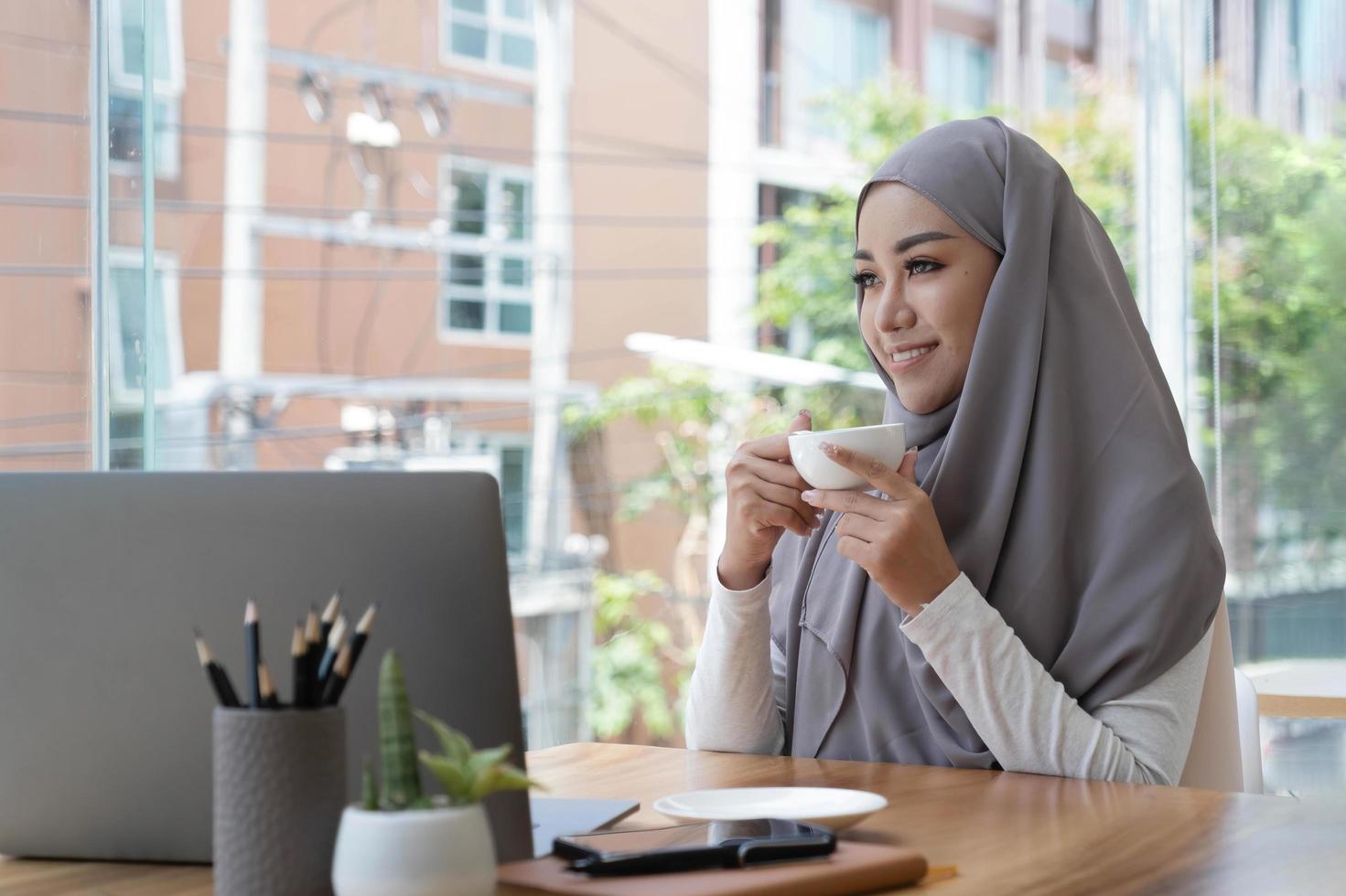 sonriente hermosa mujer de negocios musulmana bebiendo café caliente sentada en el espacio de trabajo y mirando la ventana afuera pensando en el plan de trabajo. foto