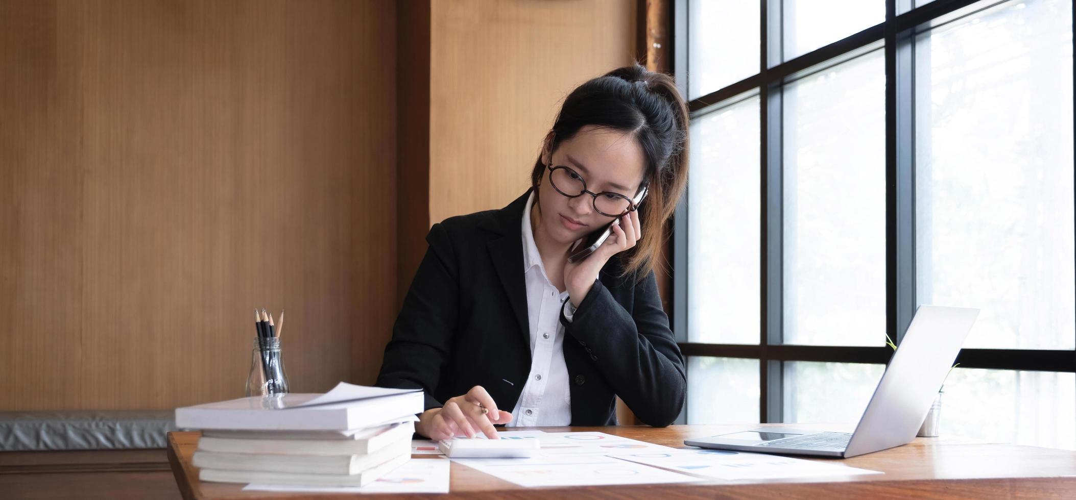 Serious asian businesswoman using laptop looking at computer talking on phone consult client sit at desk, focused female sales manager make mobile business call discuss work with customer in office photo