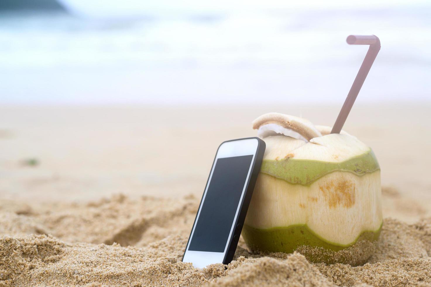 Fresh coconut with drinking straw with smartphone on the beach, travel and holidays concept. photo