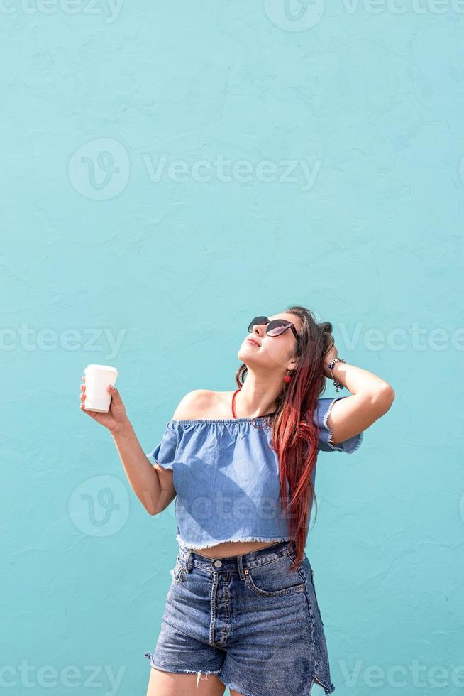 cheerful trendy woman with red hair drinking coffee dancing on blue wall background at street photo