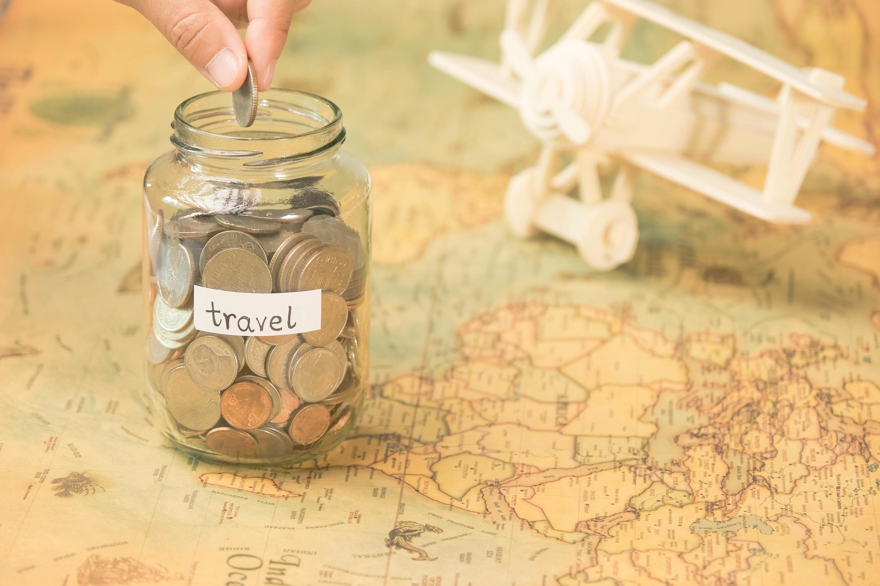 Hand putting coins in a clear jar on the table to save for travel. Glass jar  with coins and an inscription travel on the world map with wooden toy plane  on the