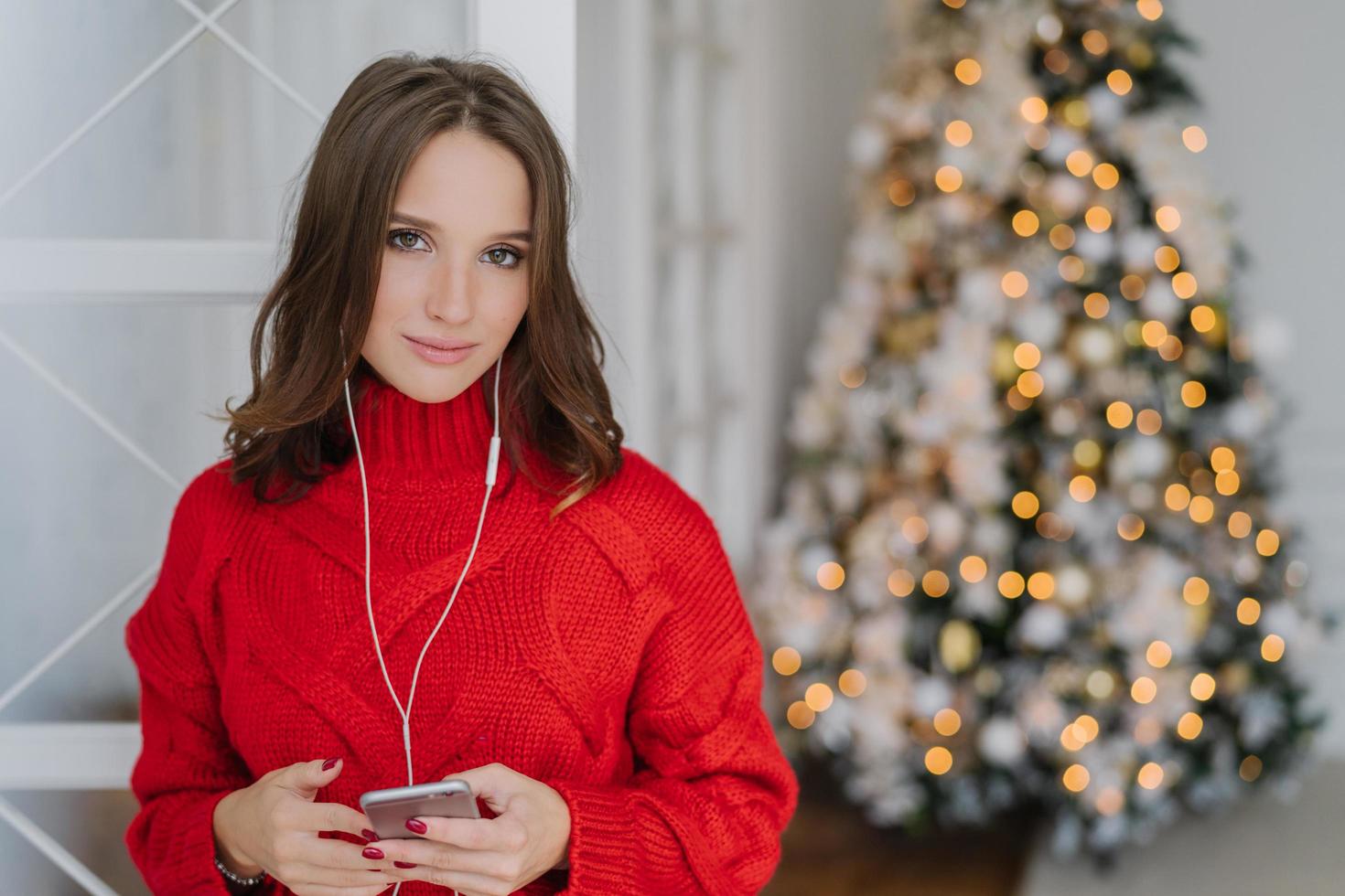 Photo of cute young woman with dark hair, dressed in knitted red sweater, holds smart phone in hands, listens radio online, connected to wifi at home, pose near beautiful New Year tree with lights