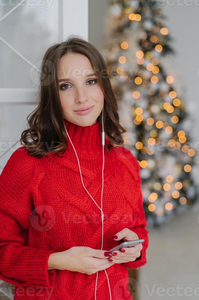 tranquila mujer encantadora y despreocupada con piel sana, vestida con suéter rojo, usa teléfono móvil y auriculares, disfruta del ambiente doméstico, pasa la Navidad en casa. concepto de personas y tecnología foto