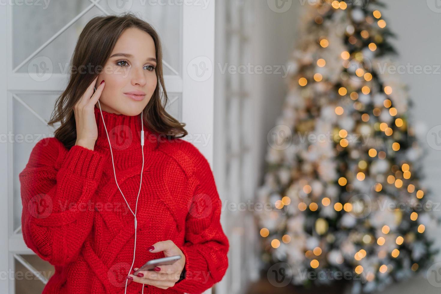 People, holidays and technology concept. Pretty woman uses mobile phone and earphones for listening music, stands at home against Christmas tree lights background with copy space for your text photo