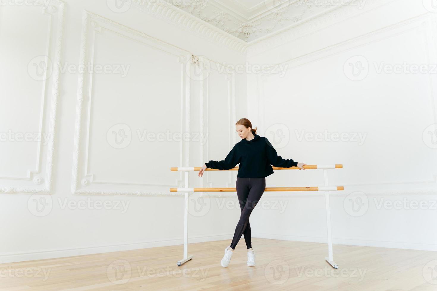 Classic ballet dancer has stretching workout, stands near ballet barres, wears black sweatshirt and leggings, does exercises in studio, focused down. Certified female instructor of choreography photo