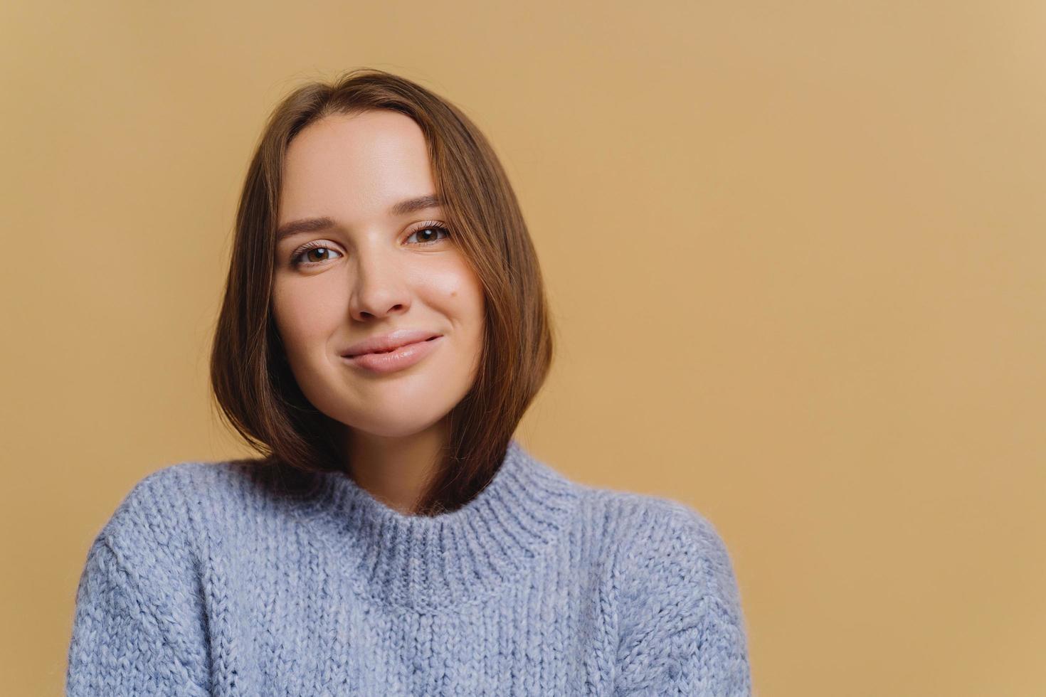 Beautiful brunette European woman with makeup, tender smile, dressed in knitwear, enjoys comfort, poses against brown background, copy space area. Lady feels comfort in new winter warm sweater photo