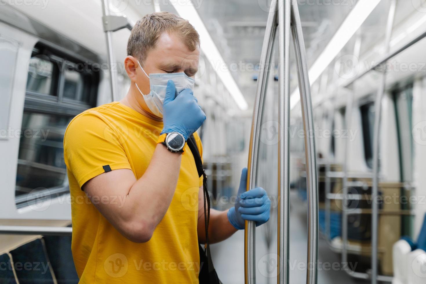 Horizontal shot of man passenger coughs and has problems with breathing, wears disposable mask and gloves, stands in public transport, metro, prevents from coronavirus. Public health solution photo