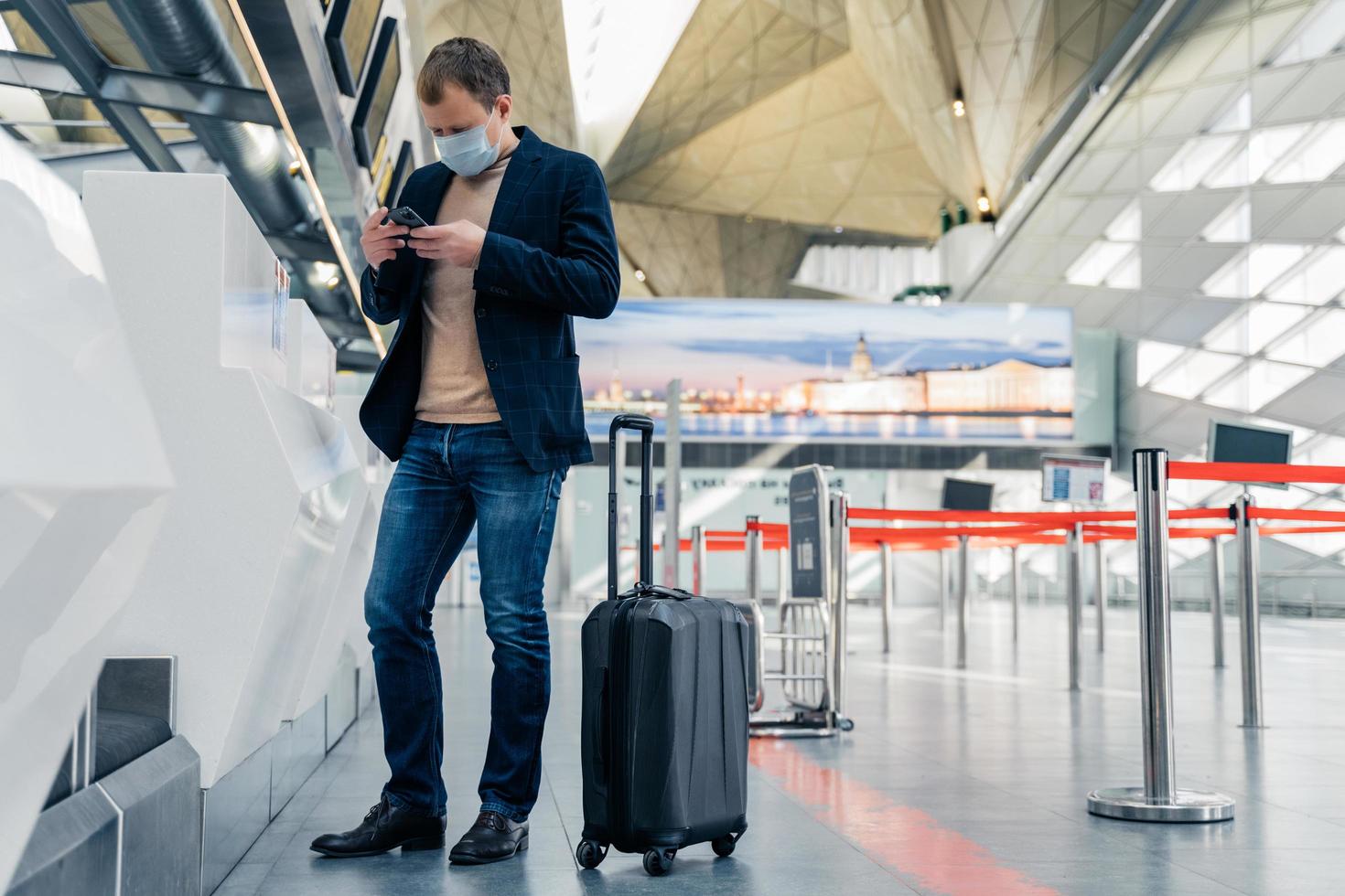 Full length shot of man passenger stands near suitcase on wheels in airport reads news online on cellular wears protective medical mask finds out about closing of countries borders because of virus photo