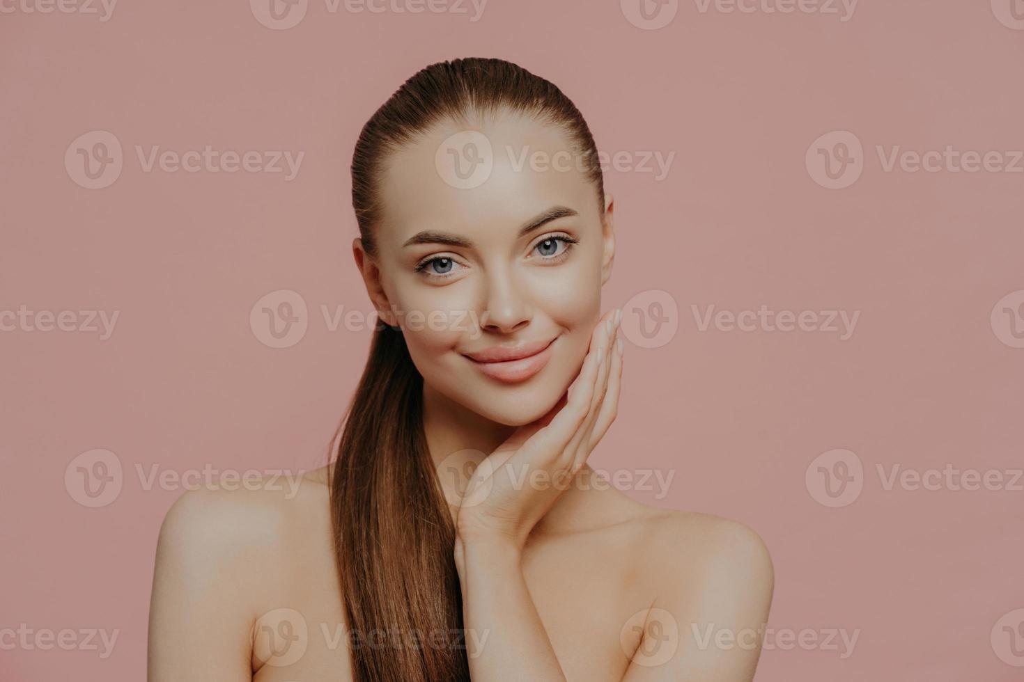 Portrait of good looking female has long straight hair combed in pony tail, stands shirtless, has clean perfect skin, satisfied with her natural beauty, poses indoor against pink background. photo