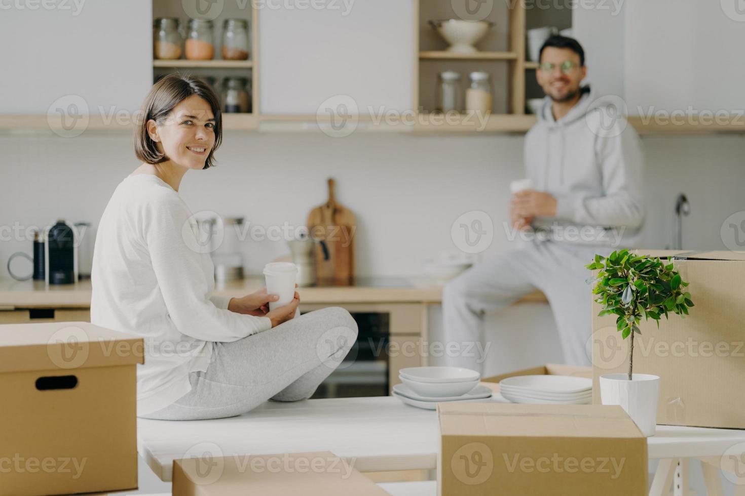 Horizontal shot of happy young woman and man sit on kitchen desk, drink coffee, have break after unpacking boxes, have pleasant talk, surrounded with plates, indoor plant in pot. Moving concept photo