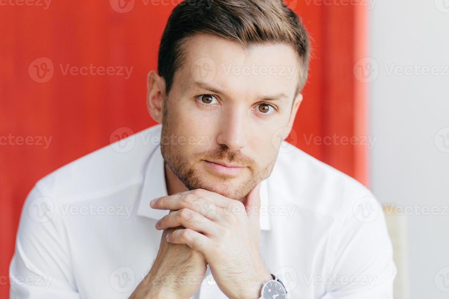 Indoor shot of pensive young male office worker keeps hand together under chin, wears white shirt, looks directly at camera, contemplates about business meeting, isolated over ed background. photo