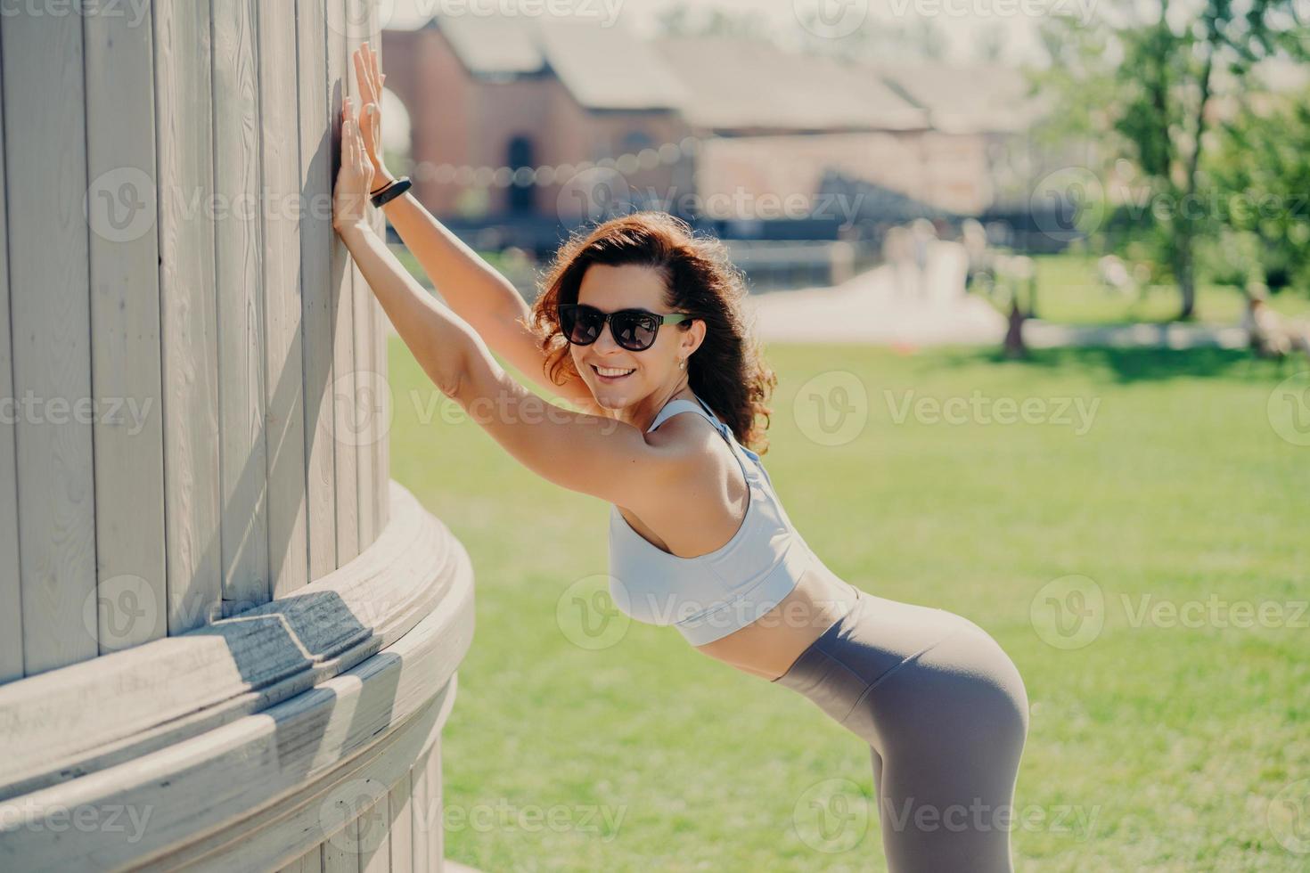 Slim woman with perfect body shapes leans with hands at something smiles positively dressed in top and leggings poses outdoor has fitness training during summer time. People and exercising concept photo