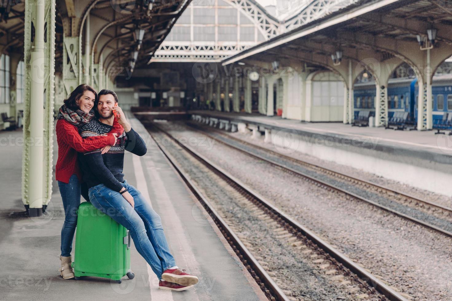 People, travelling, relationship concept. Happy unshaven husband and wife embrace at railway station, going to have trip, have vacation. Handsome man speaks on smart phone, recieves hug girlfriend photo