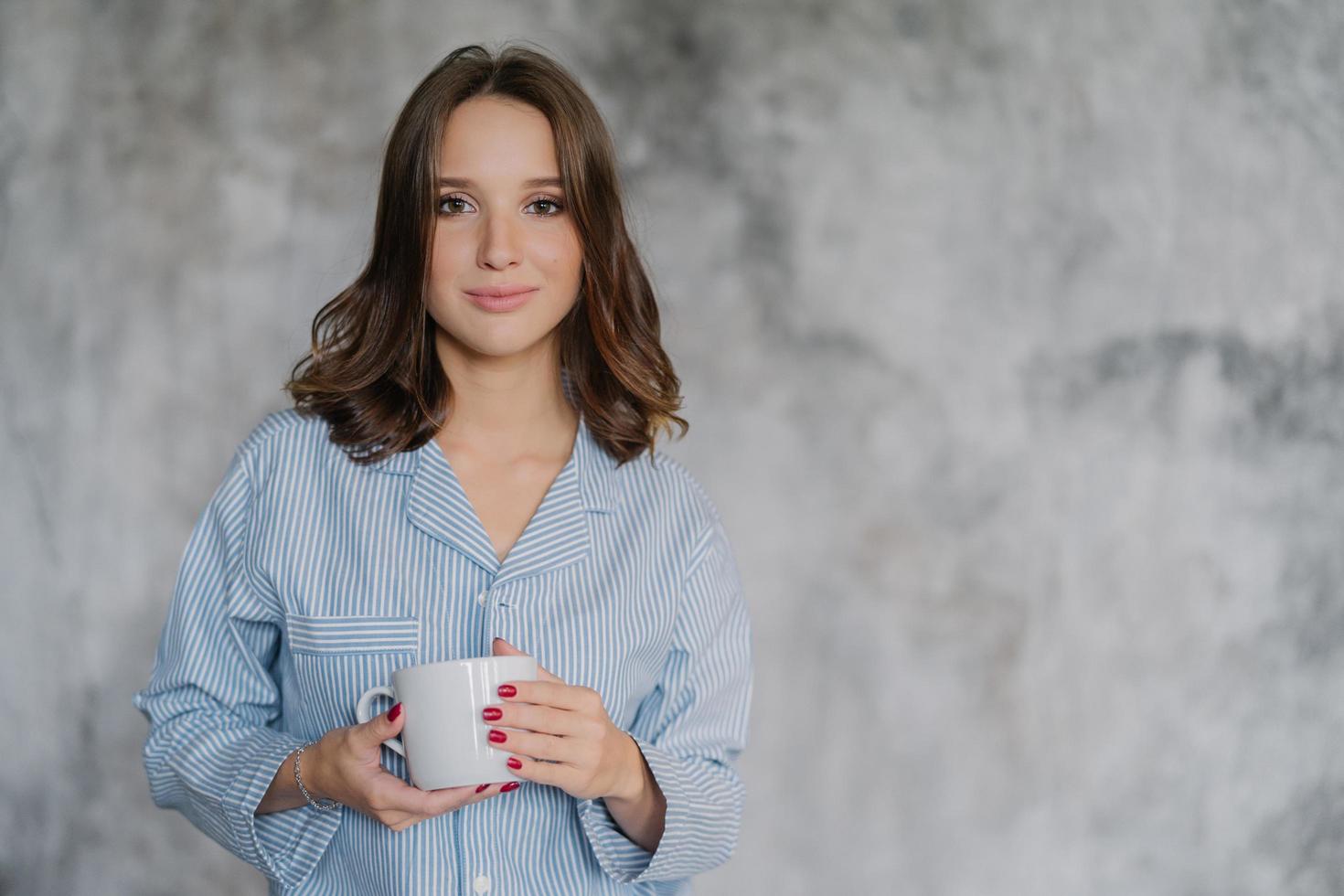 Attractive woman dressed in nightwear, holds white mug with coffee or tea, has morning drink, poses indoor against blurred background with copy space for your advertisement or promotional content photo