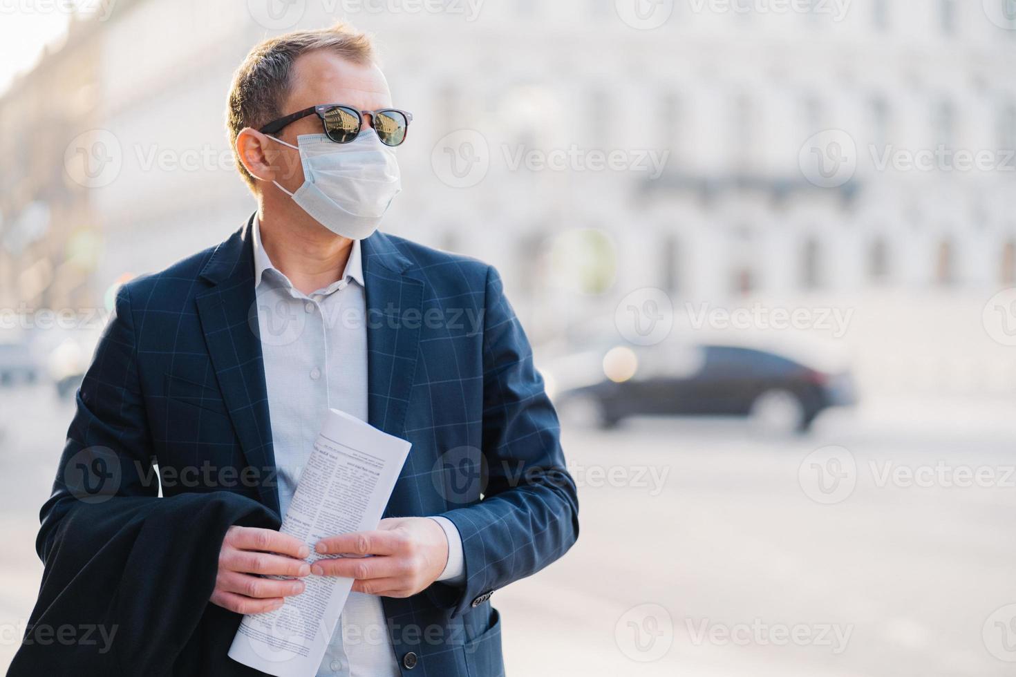 Covid 19 Man Worker Dressed In Formal Wear Holds Newspaper In Hands Poses At Street Wears Surgical Mask Avoids Crowded Street Not To Catch Virus Poses Against Blurred Background With Transport Stock