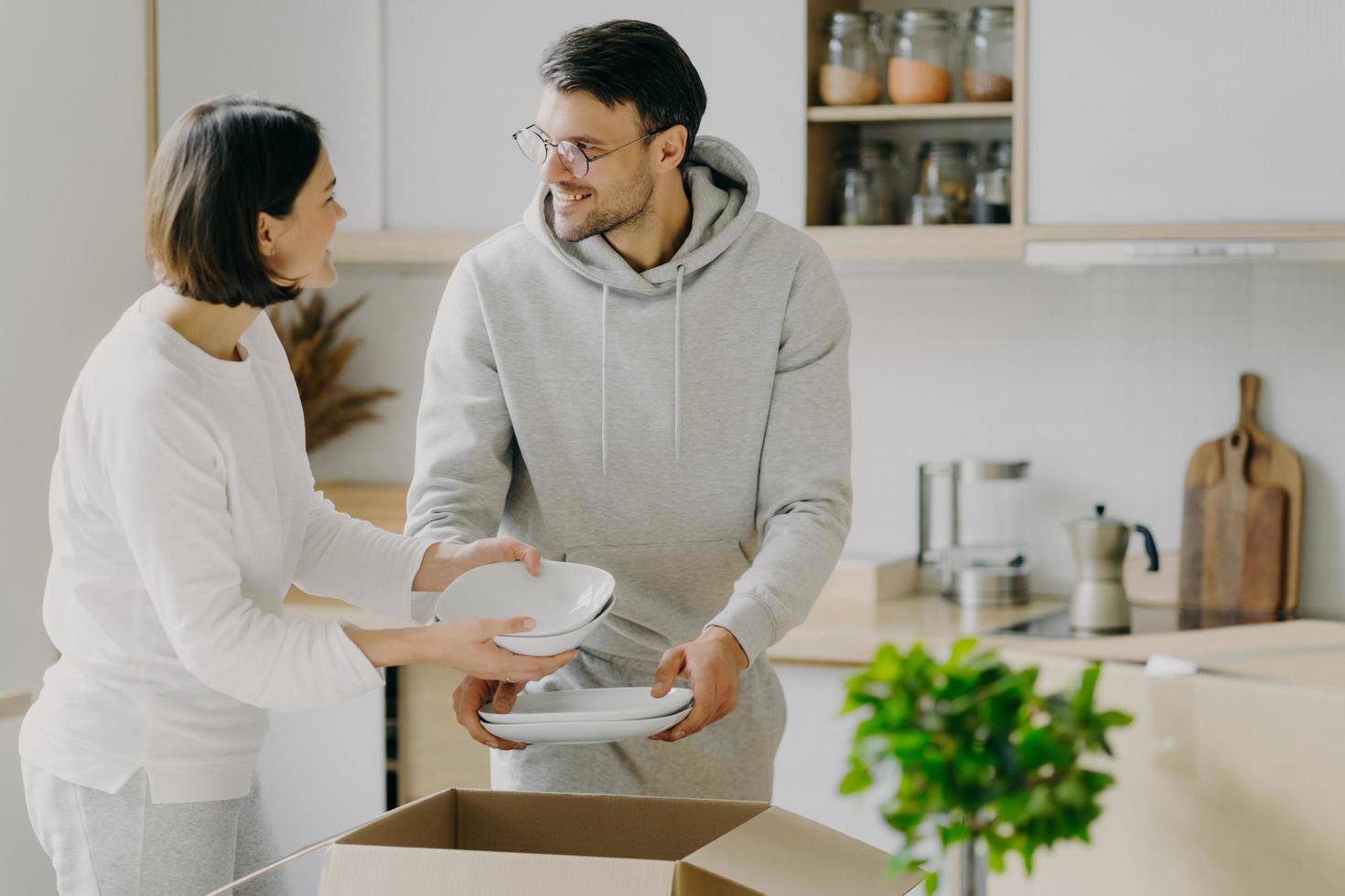 Delighted housewife and her husband unpack cardboard boxes with new tableware, carry plates, move into new apartment, look gladfully at each other, pose agaist kitchen interior. Relocation concept photo