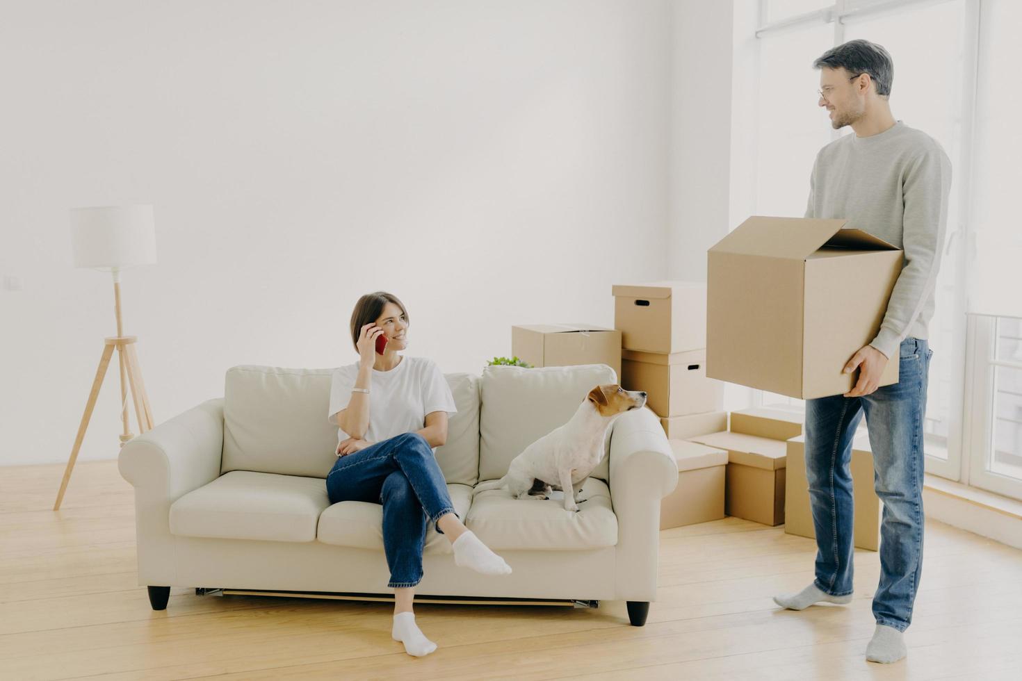 Woman and man real estate renters pose in empty living room, female sits on couch in middle of room, has phone conversation, share impressions after moving in new apartment, male carries boxes photo