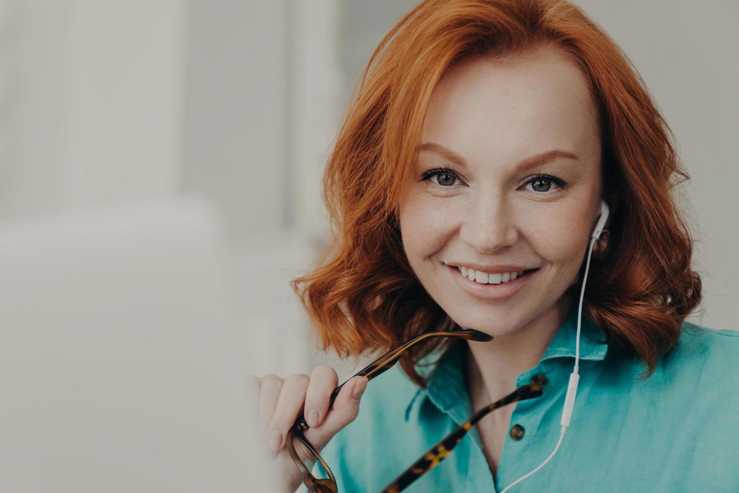 Portrait of beautiful redhead professional female teaches conducts lessons online, waits for starting video conference with pupils, uses earphones and notebook, smiles positively, holds spectacles photo