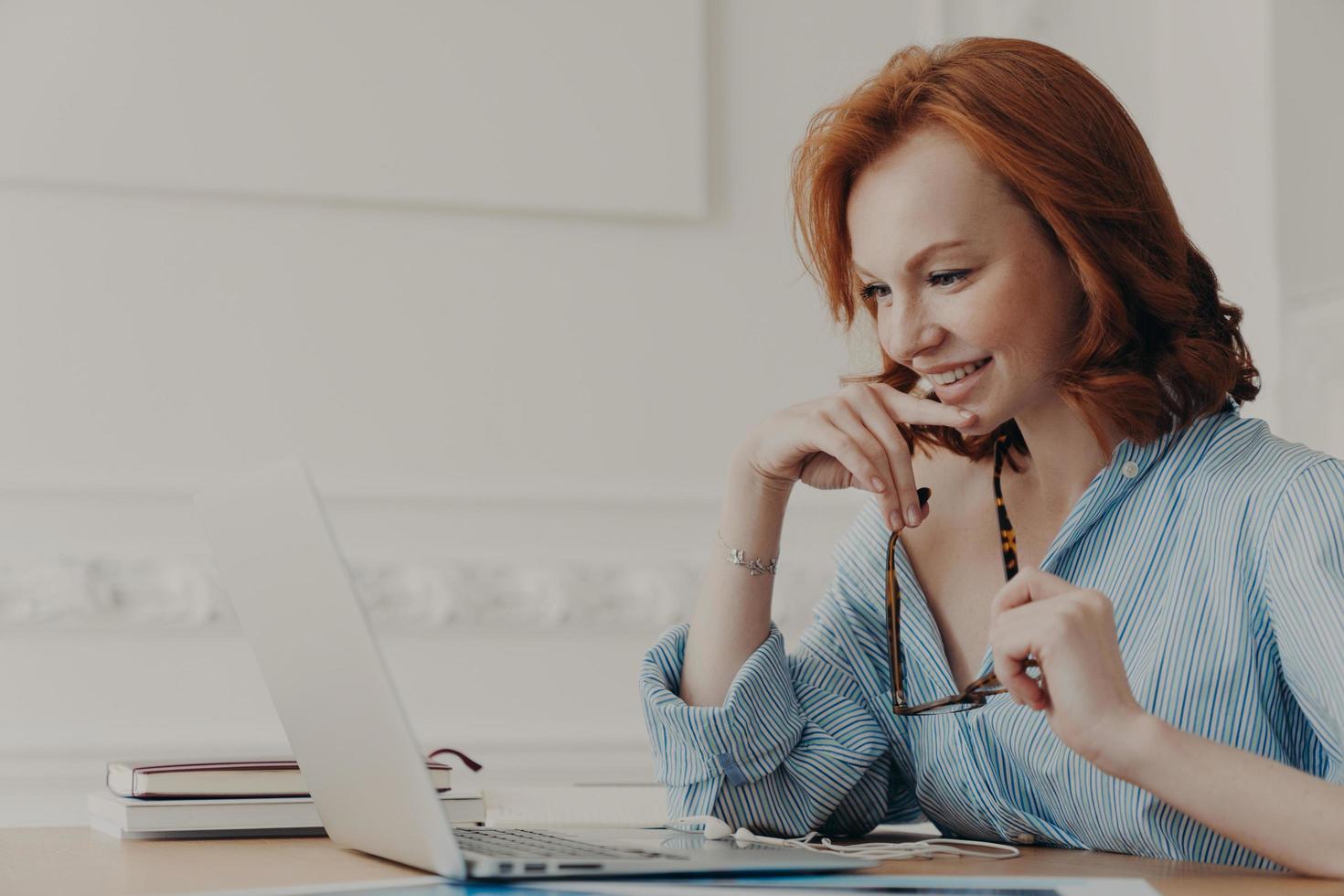 Pleased female model poses at coworking space, holds spectacles, smiles positively, uses wireless internet connection, reads information, has online communication, works on creating web project photo