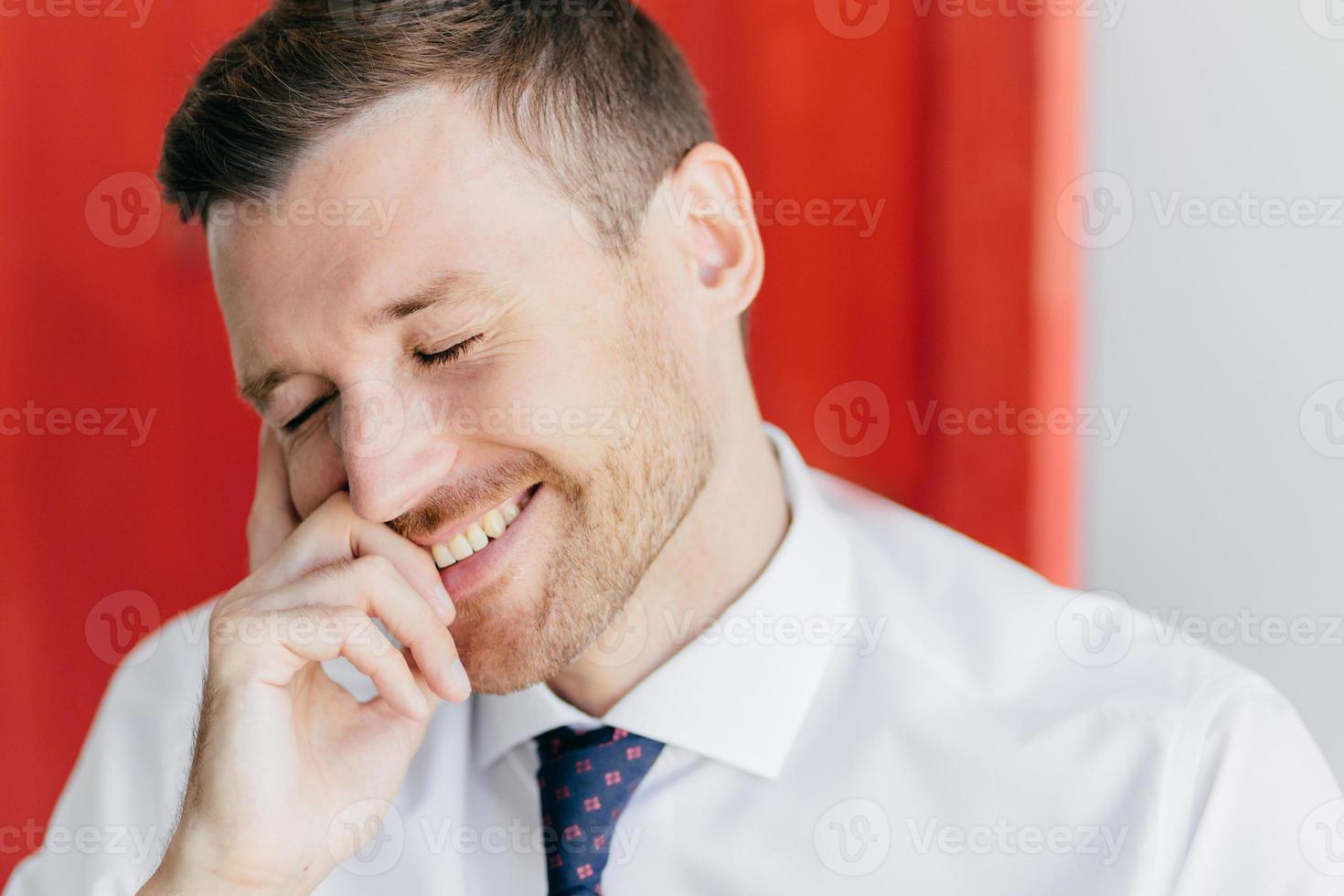Joyful unshaven male with happy expression, giggles at funny joke, closes eyes with pleasure, dressed in elegant white shirt, recieves pleasant news, poses against red wall. Positive emotions concept photo